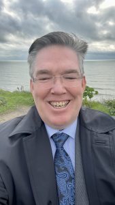 Man smiling outdoors by the seaside wearing a suit and tie under cloudy skies
